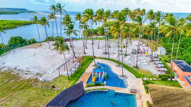 Vista aérea de um resort de praia tropical com palmeiras, piscinas, areia branca e lagoa azul-turquesa.