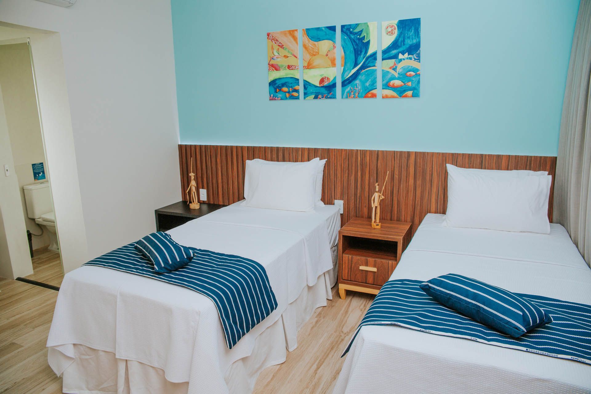 Duas camas de solteiro em um quarto iluminado com paredes azuis, lençóis brancos e uma pintura de praia acima das cabeceiras.