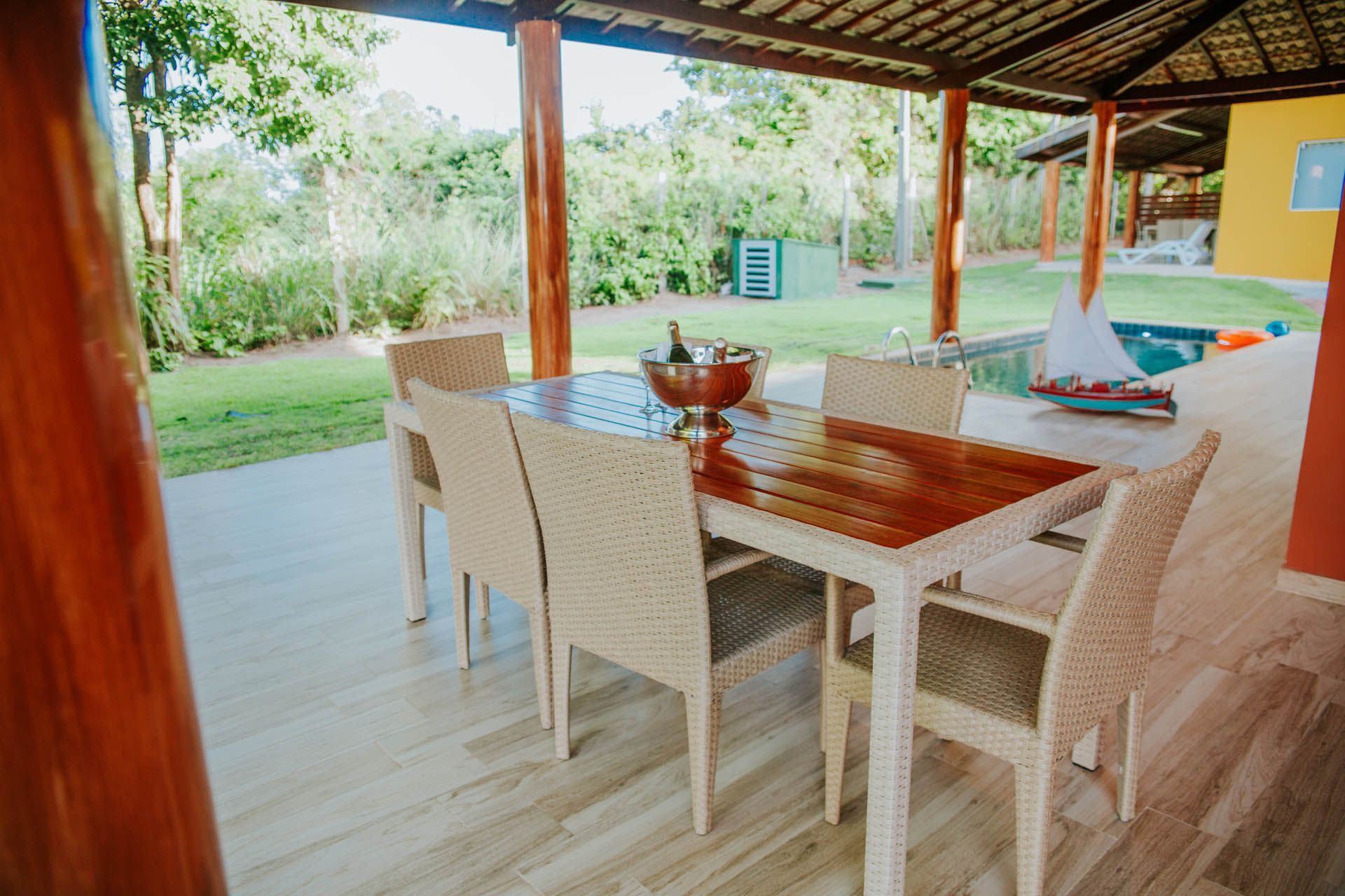 Área de jantar coberta no pátio, com mesa e cadeiras de vime, com vista para um jardim gramado.