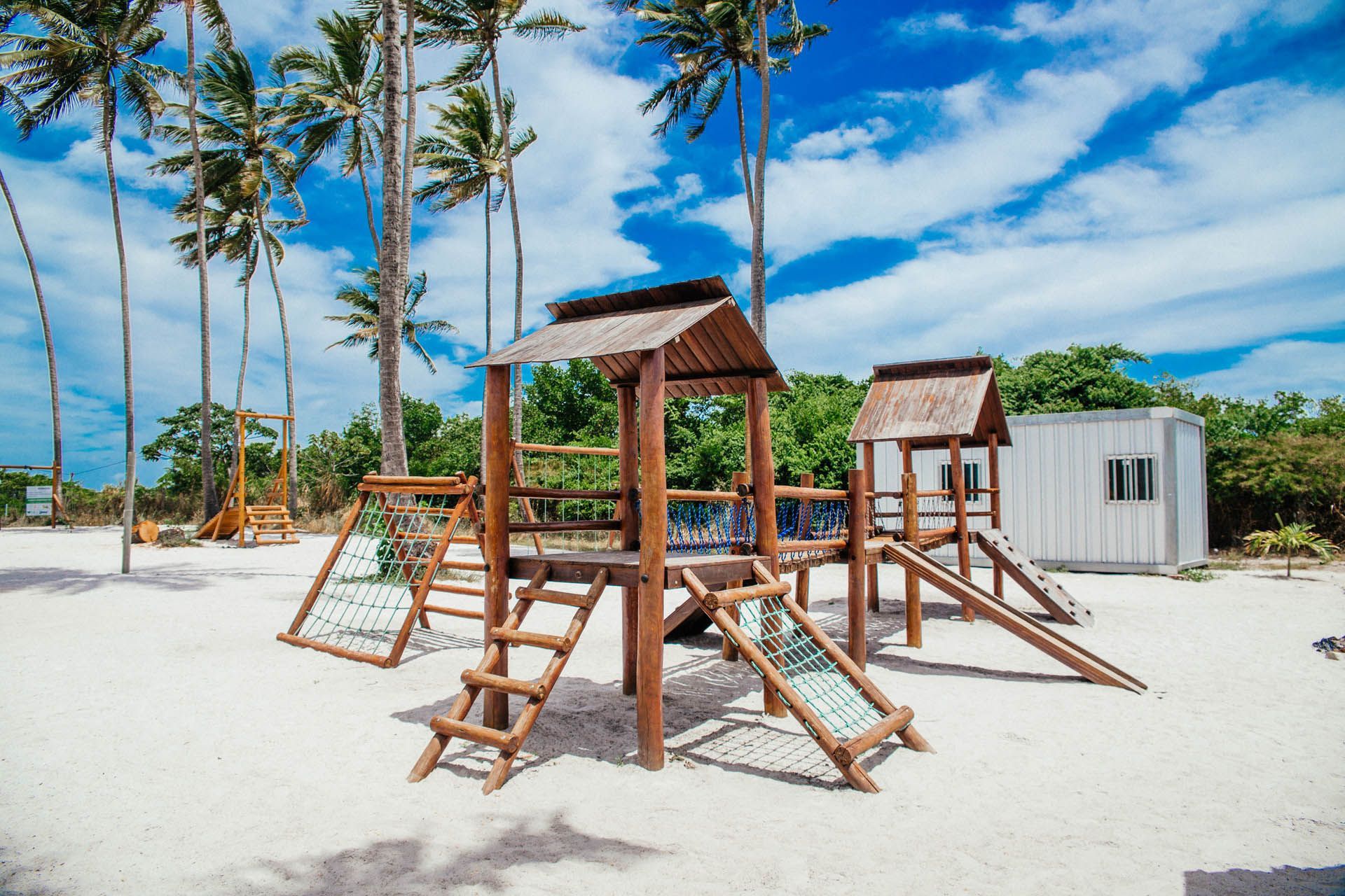 Parque infantil de madeira na praia, com areia branca, palmeiras e céu azul.