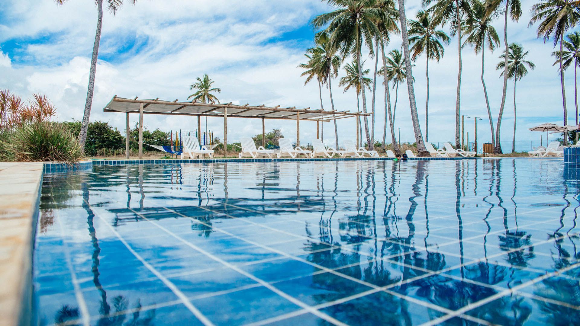 Piscina infinita com azulejos azuis, palmeiras e vista para o mar sob um céu parcialmente nublado.