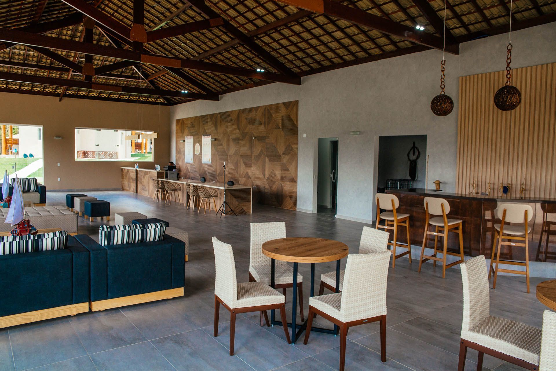 Interior de restaurante ao ar livre com mesas e cadeiras de madeira, balcão de bar e cobertura de palha.
