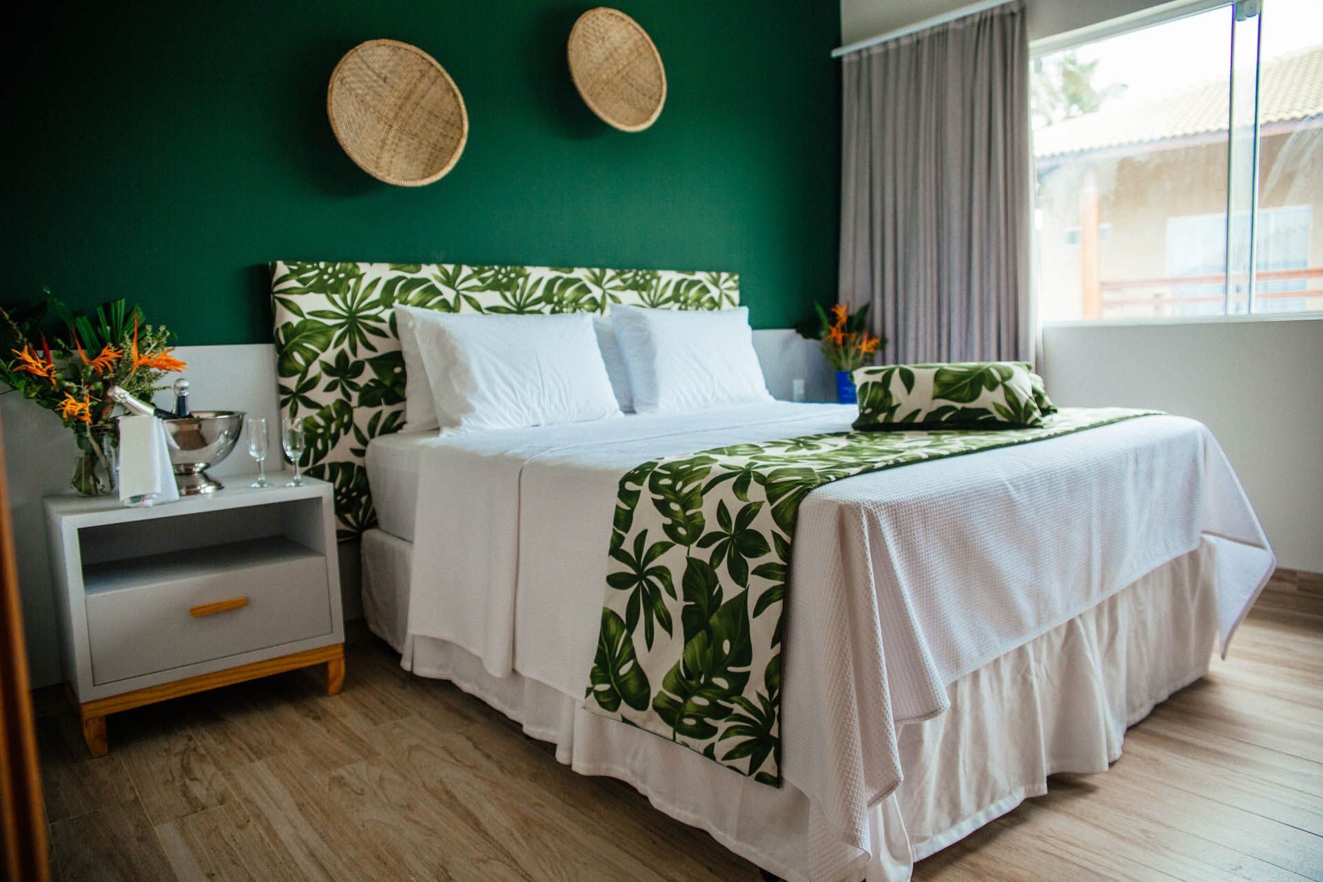 Quarto luminoso com parede de destaque verde, cama branca e decoração tropical com folhagens.