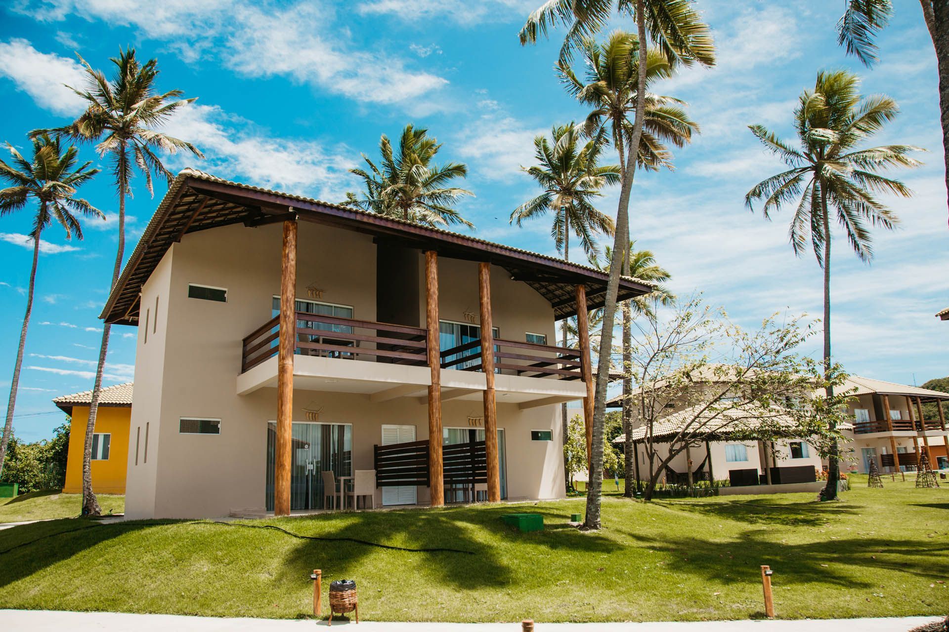 Casa tropical de dois andares com varanda, palmeiras e gramado verde sob um céu azul ensolarado.