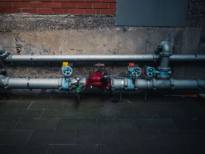 Industrial pipes and valves against a brick wall on a wet pavement, with a red central valve assembly