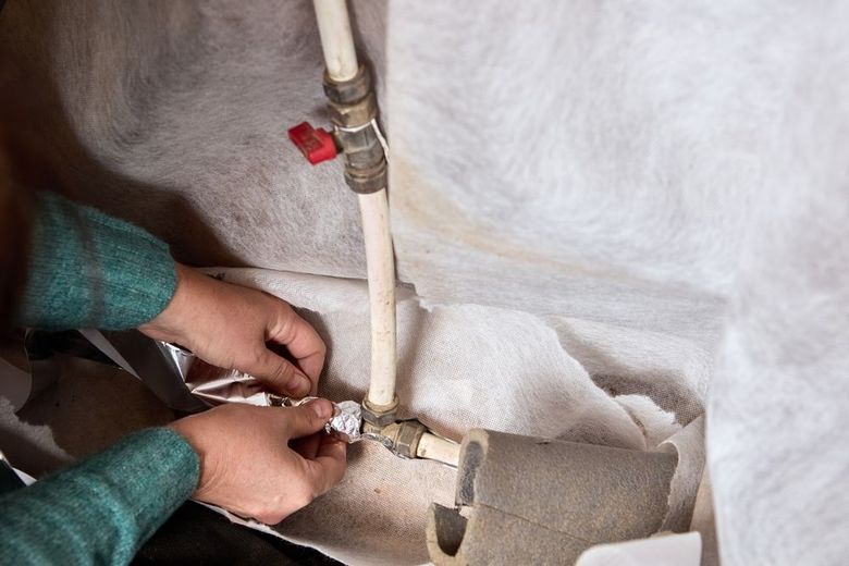 Hands connect a gas line valve under a gray insulation cover, with a red shutoff lever visible.