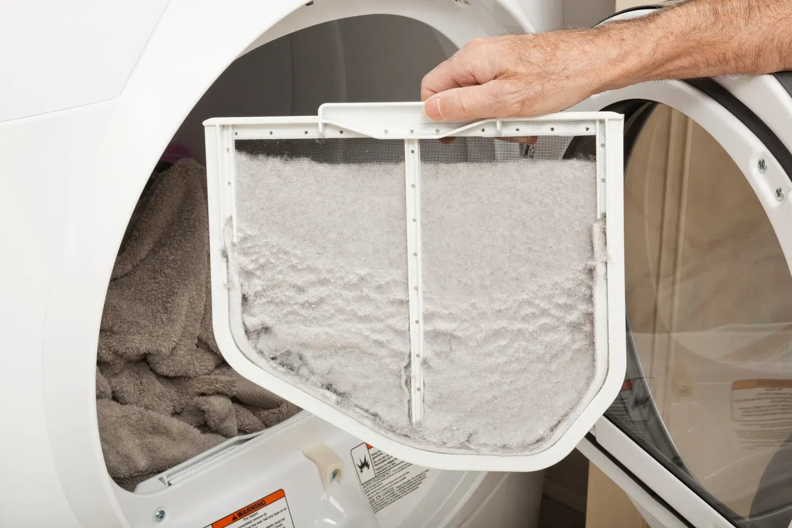 Person empties a lint trap covered in white dryer lint from a front-loading dryer