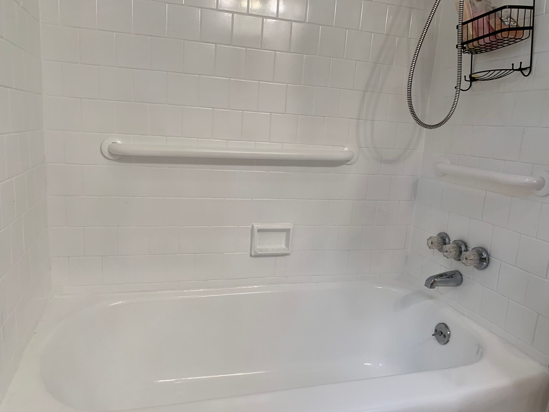 White tiled bathtub and shower area with grab bars, a handheld showerhead, and soap holder