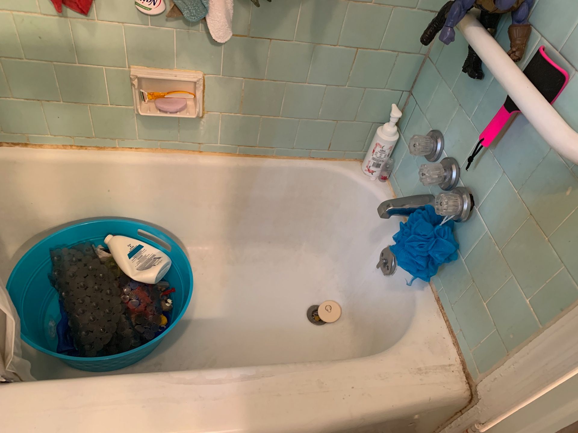 Cluttered bathtub with a blue basin of laundry, soap, and a bath sponge in a tiled bathroom.
