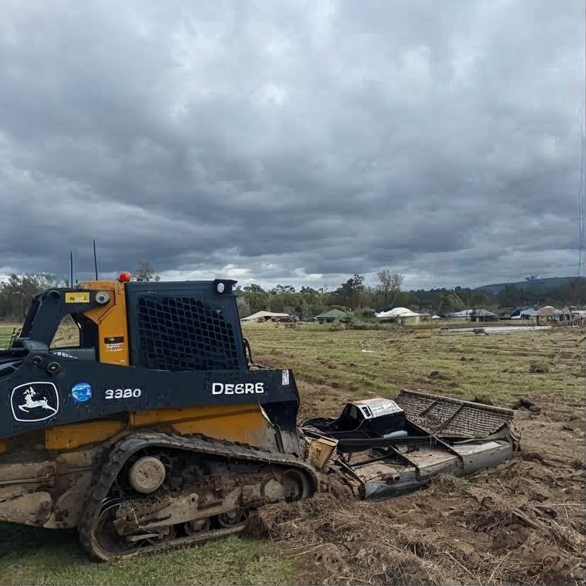 John Deere bulldozer in a muddy field under dark storm clouds, with a damaged attachment on the ground