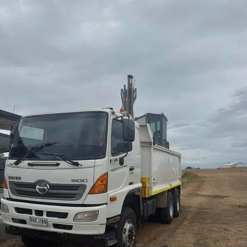 White dump truck parked on a dirt road under a cloudy sky, with a rural landscape behind it