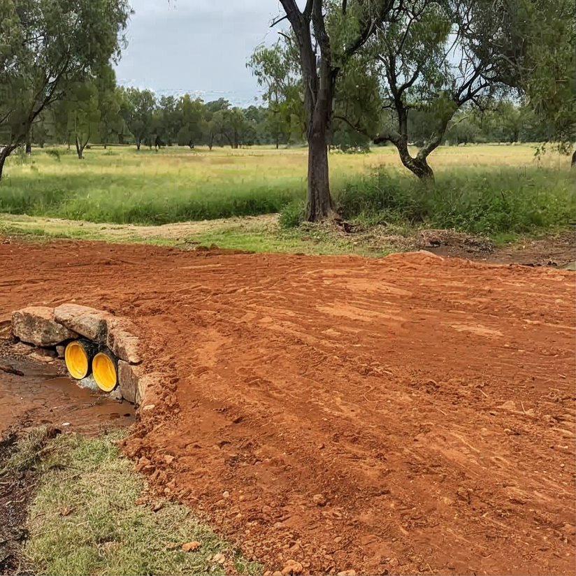 Red dirt road beside grassy savanna with trees and a stone culvert pipe