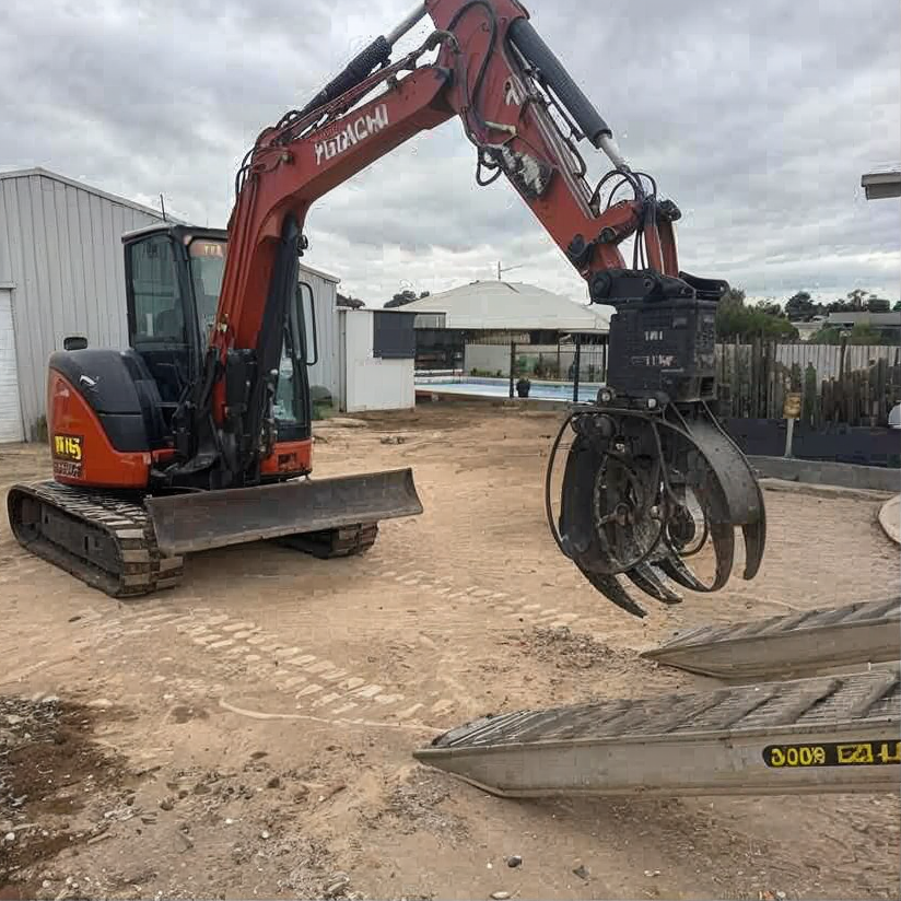 Orange excavator with grapple attachment parked on a dirt lot near buildings and a fence