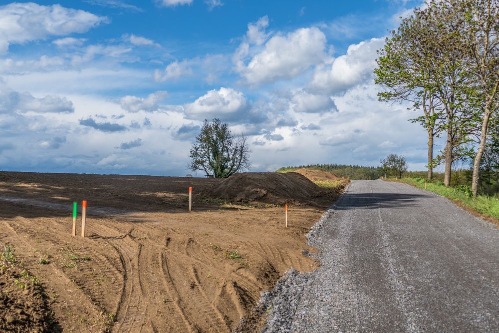 Gravel road beside a cleared dirt field under a cloudy blue sky, with trees along the right side.