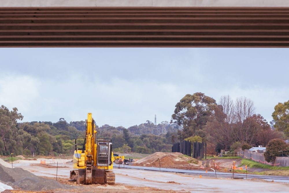 Yellow excavator on a muddy construction site with dirt piles and trees under a cloudy sky