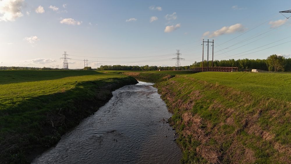 Narrow stream flowing through grassy fields under a clear sky, with power lines in the distance