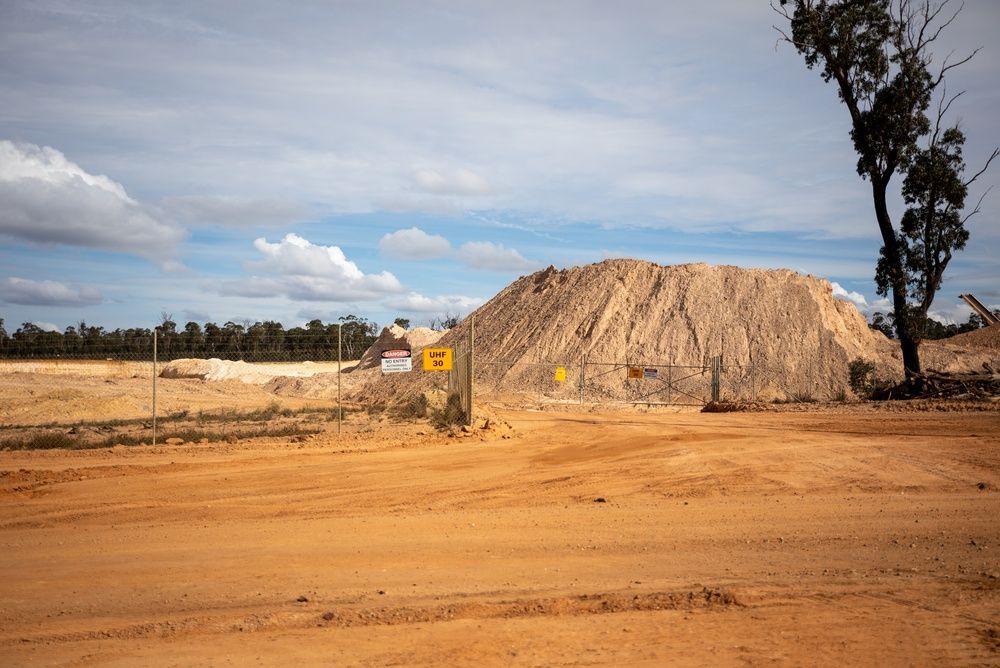 A large dirt mound on a construction site under a cloudy sky, with a sign and a tree nearby.