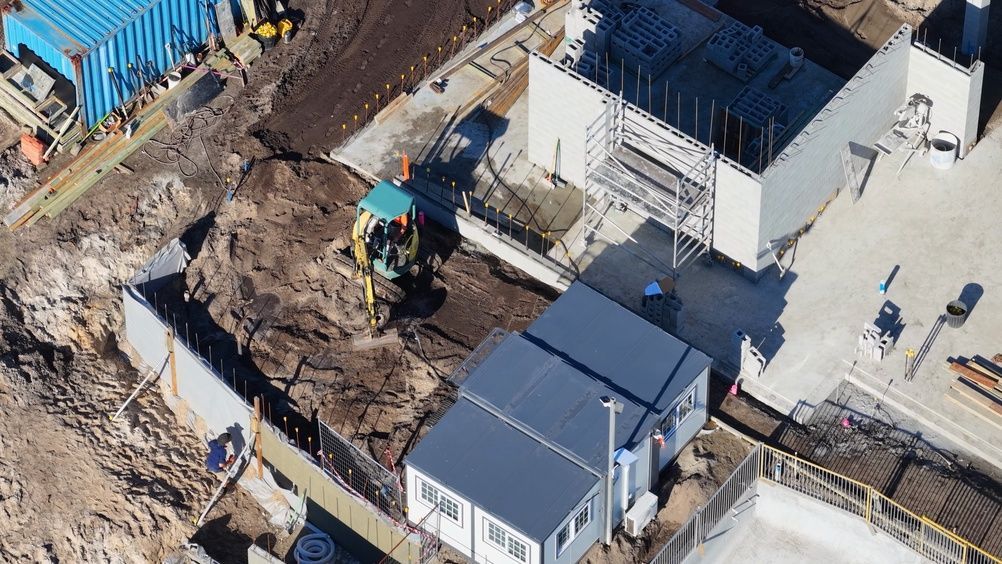 Aerial view of a construction site with an excavator digging beside white portable buildings and a fenced area.