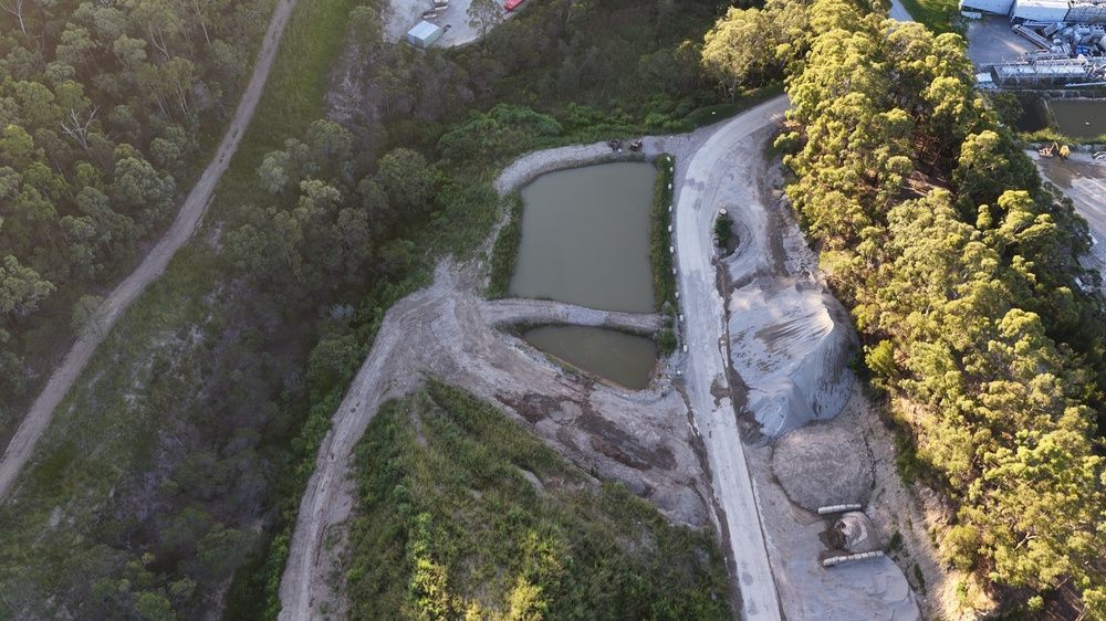 Aerial view of a small green pond beside a gravel road and rocky hillside with trees
