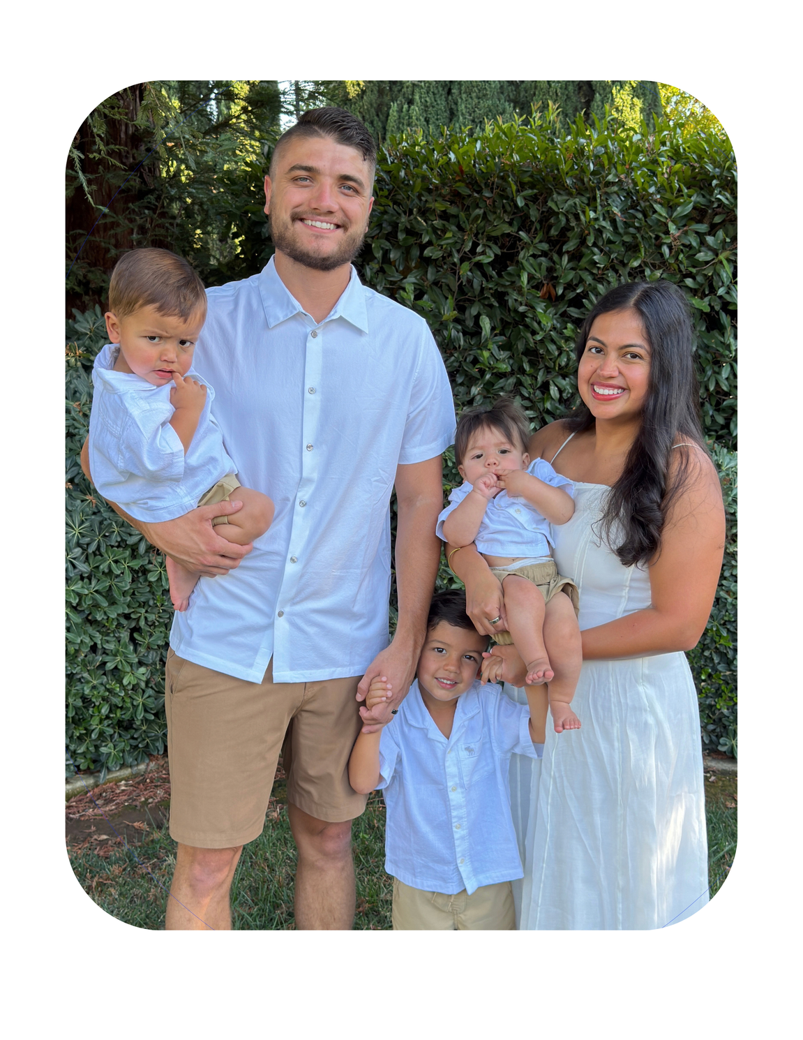 Family of four posing outdoors in front of green hedges, dressed in light blue and white outfits.