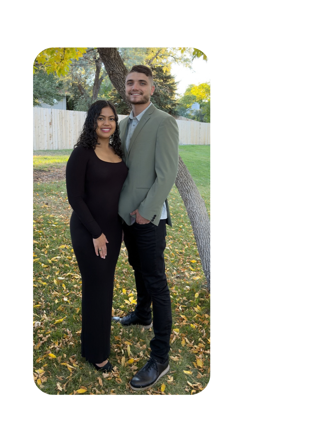 Two people standing together outdoors on a grassy path, smiling, with autumn trees and a white fence behind them.