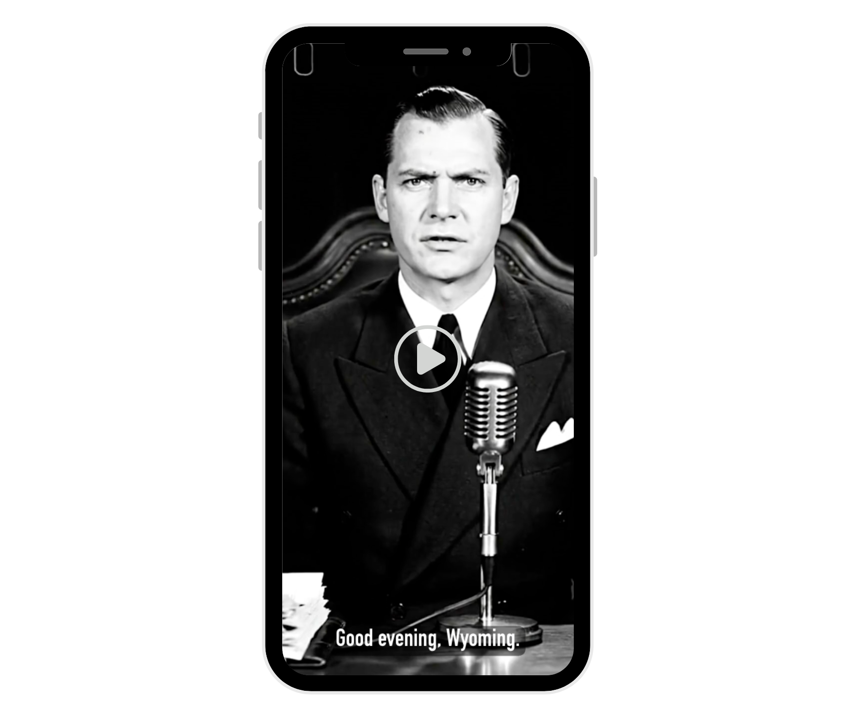 A black-and-white image of a man in a suit at a microphone, shown on a smartphone screen, with