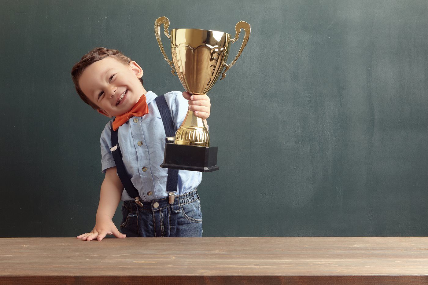 Young boy wearing suspenders and a bow tie is smiling and holding a large trophy.
