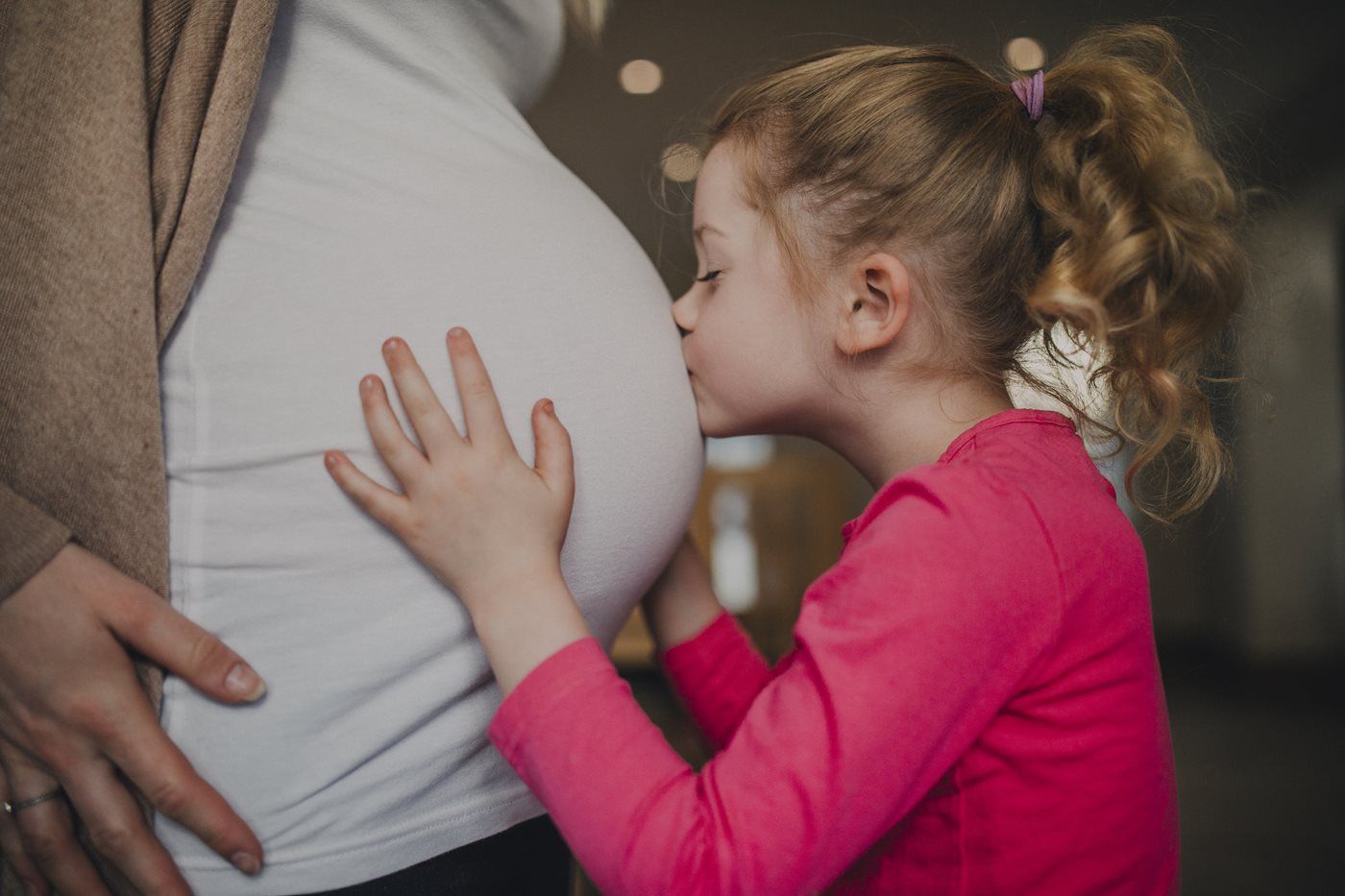 A young child in a pink shirt gently kisses and rests hands on a pregnant belly.