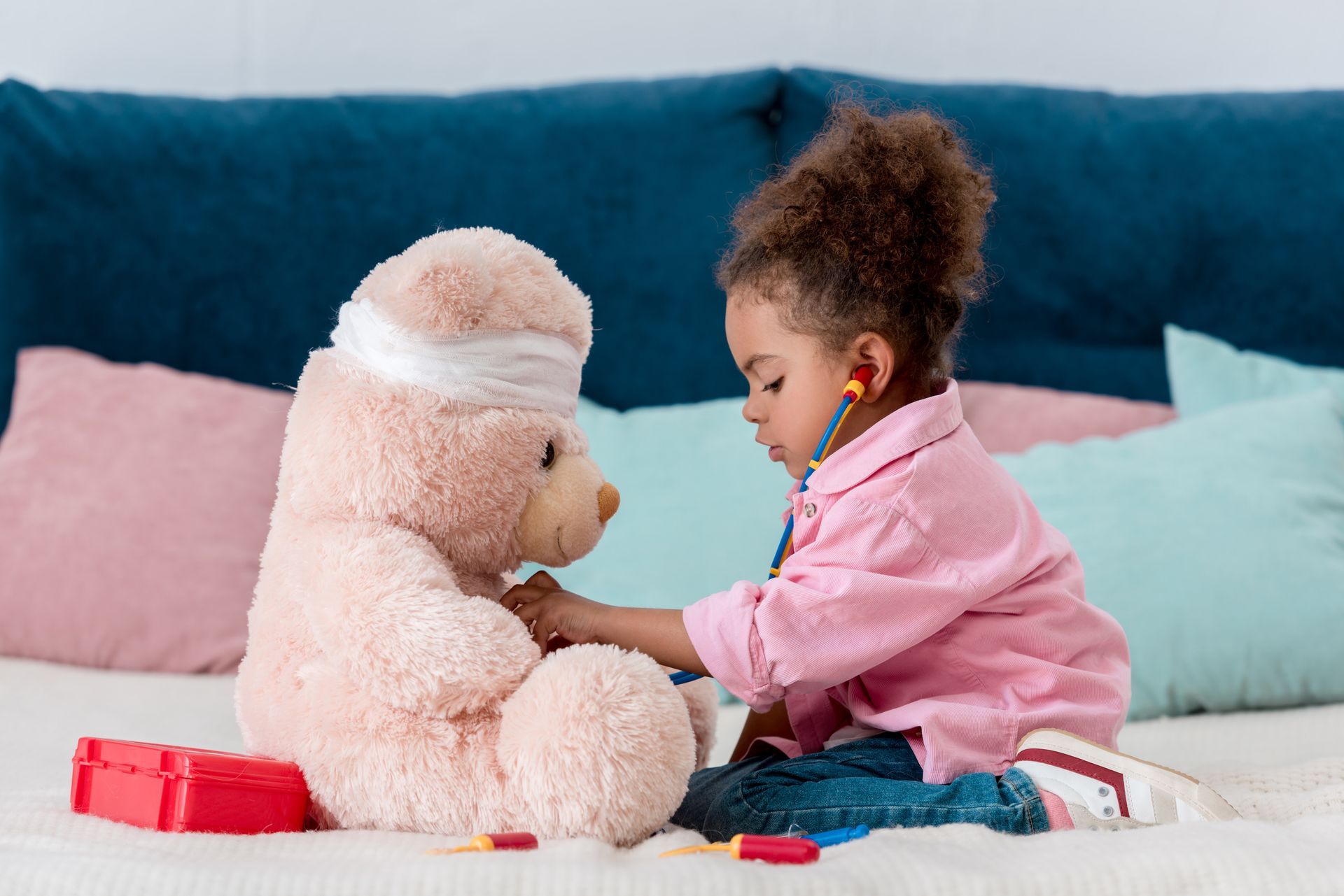 A child playing doctor with a bandage-wrapped teddy bear on a bed, using a toy stethoscope.