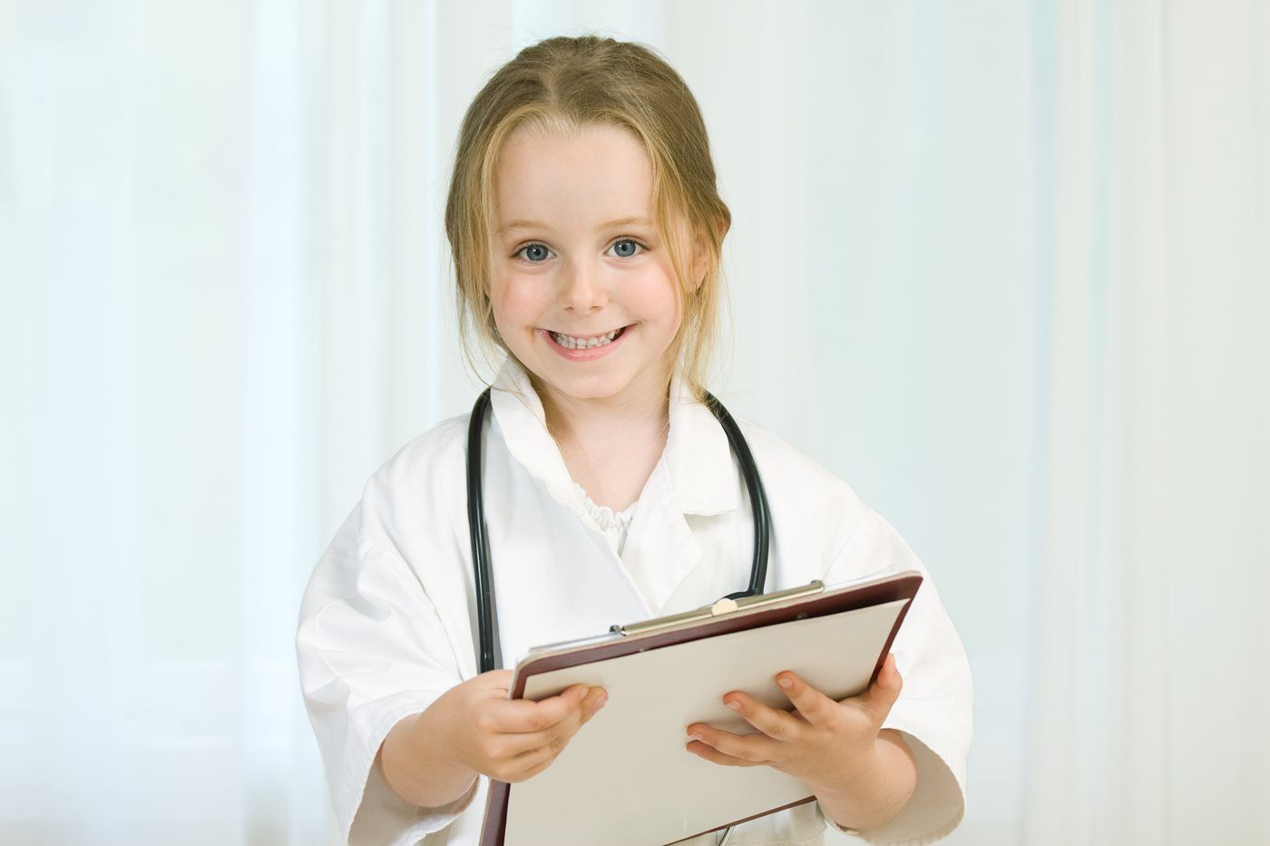 A smiling child dressed in a white lab coat with a stethoscope around their neck, holding a clipboard.