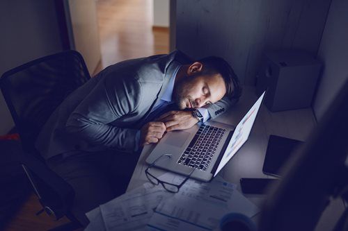 A person in business attire sleeping at their desk with a laptop, glasses, and papers in a dimly lit office.