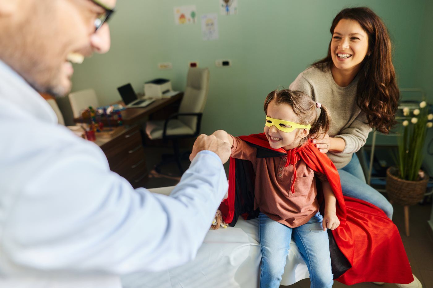 A person in a light blue shirt fist-bumps a child wearing a superhero cape and mask, while a smiling adult sits nearby.