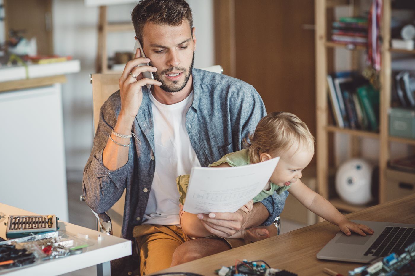 Man on phone holds toddler while working from home.