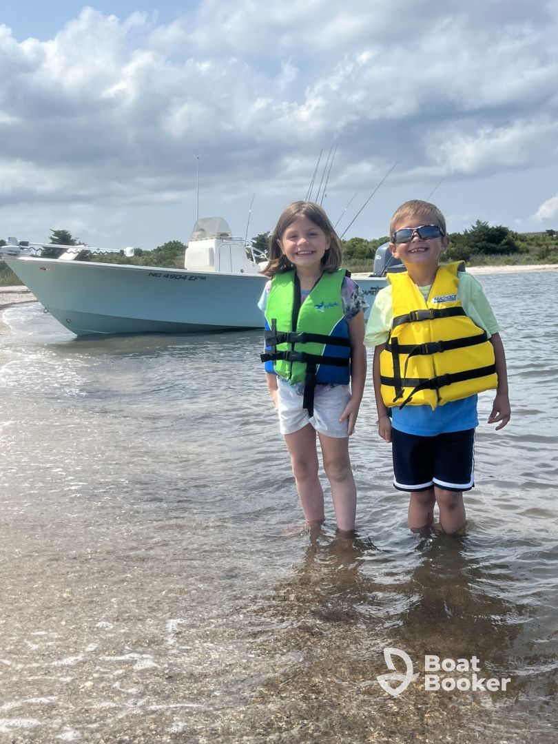 Two children in life jackets standing in shallow water beside a boat on a cloudy beach