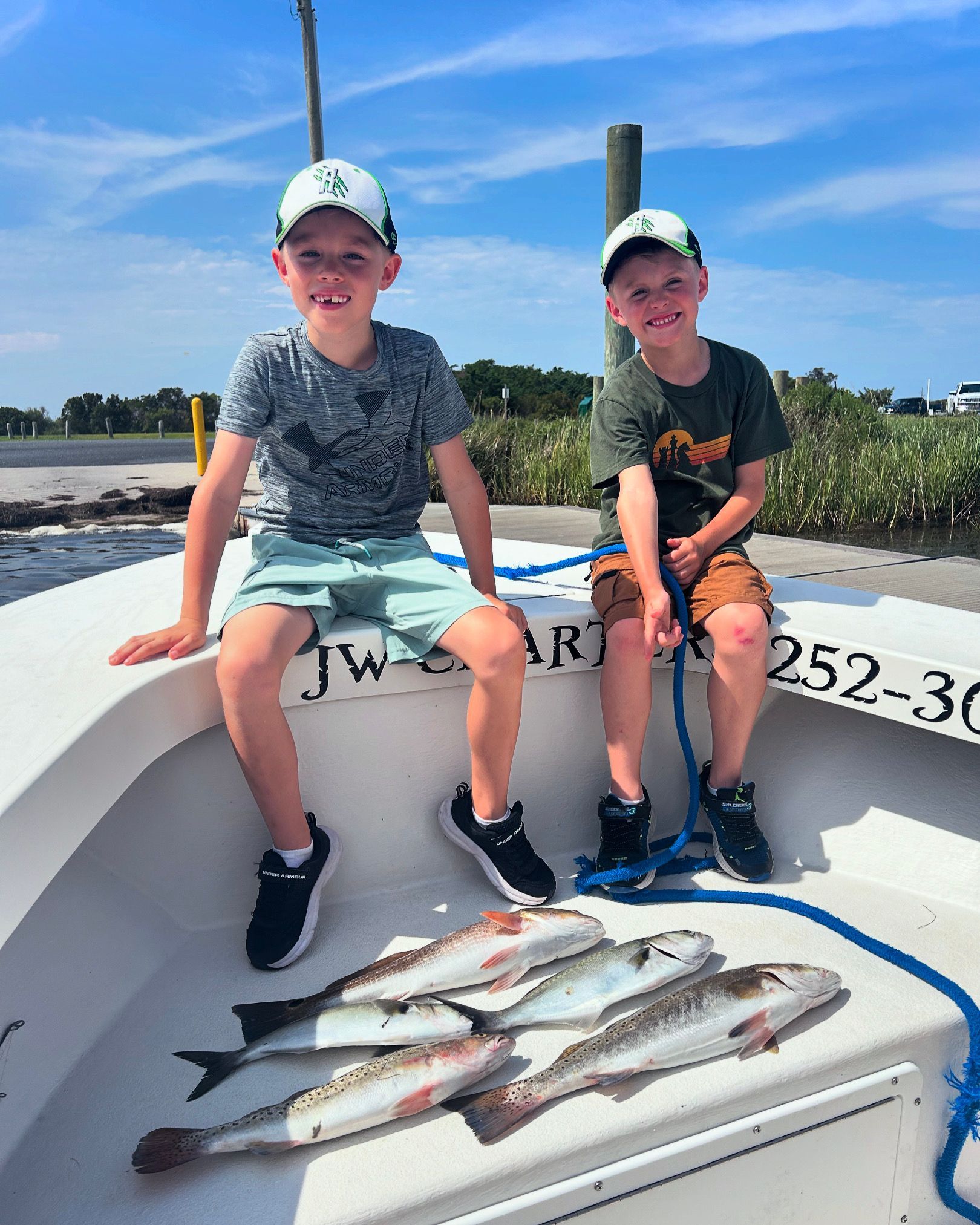 Two children sit on a boat holding fresh-caught fish on deck under a blue sky.
