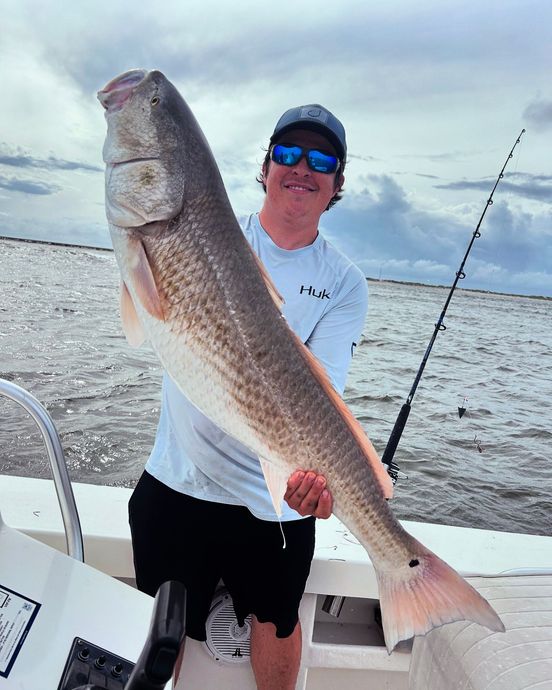 Person on a boat holding a large fish, with fishing rod and ocean in the background.