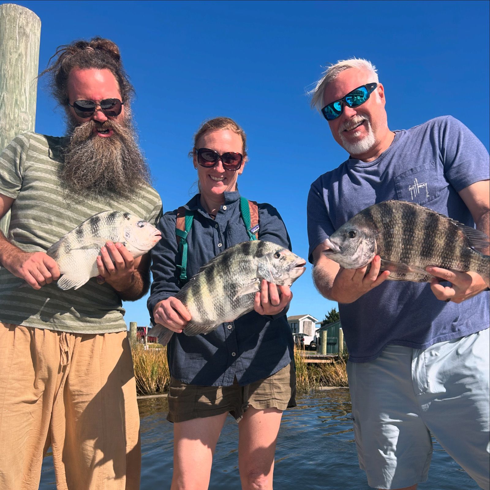 Three people holding fish by the water on a sunny day