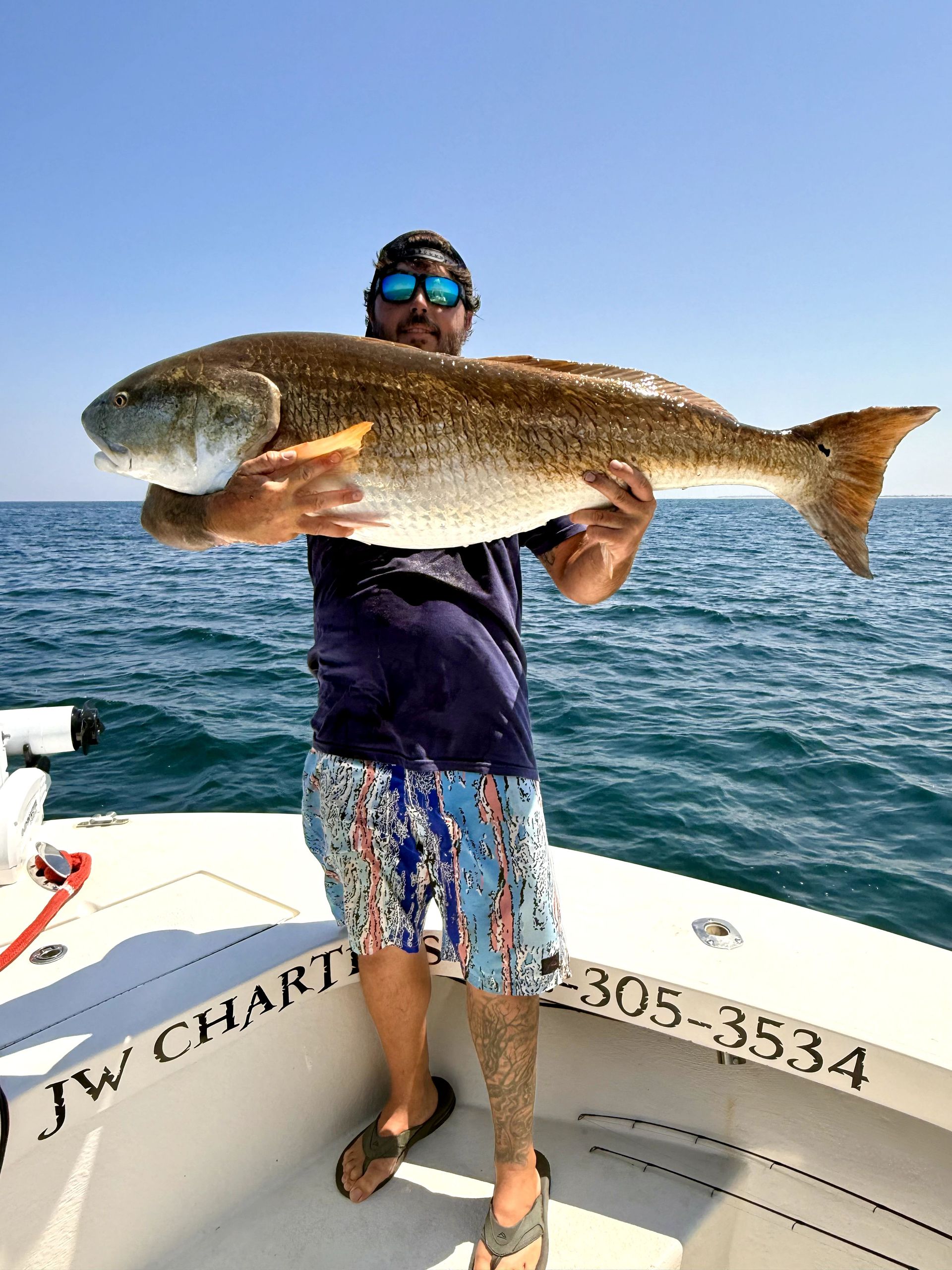 Person on a boat holding a large fish over the ocean, wearing sunglasses and patterned shorts.