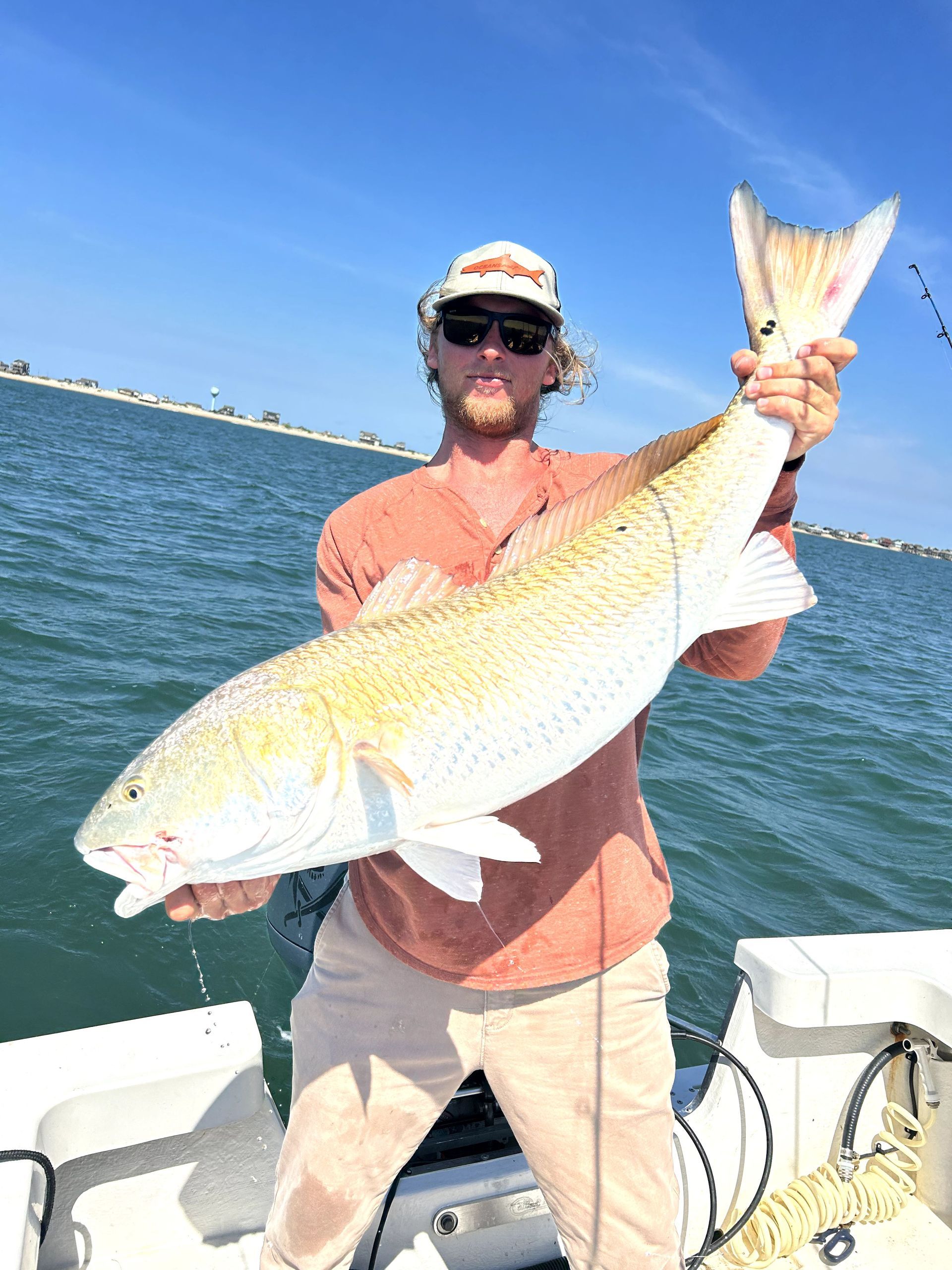 Man holding a large fish on a boat, with blue water and clear sky in the background