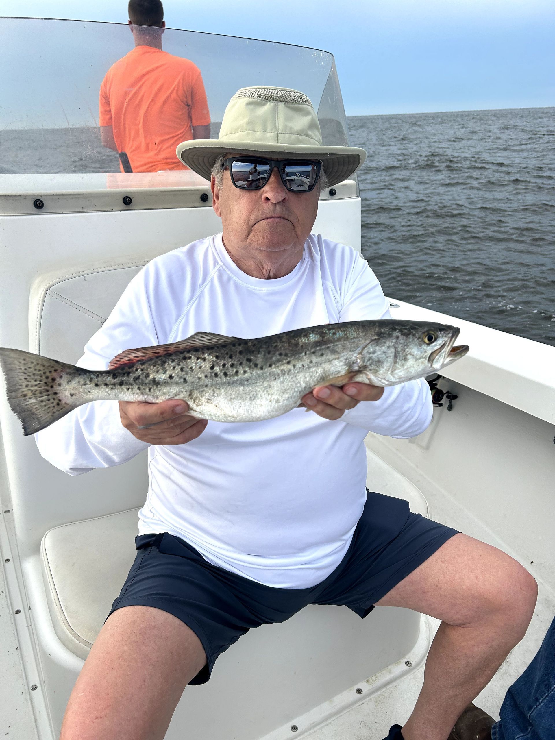 Man seated on a boat holding a large fish, wearing a white shirt, hat, and sunglasses.