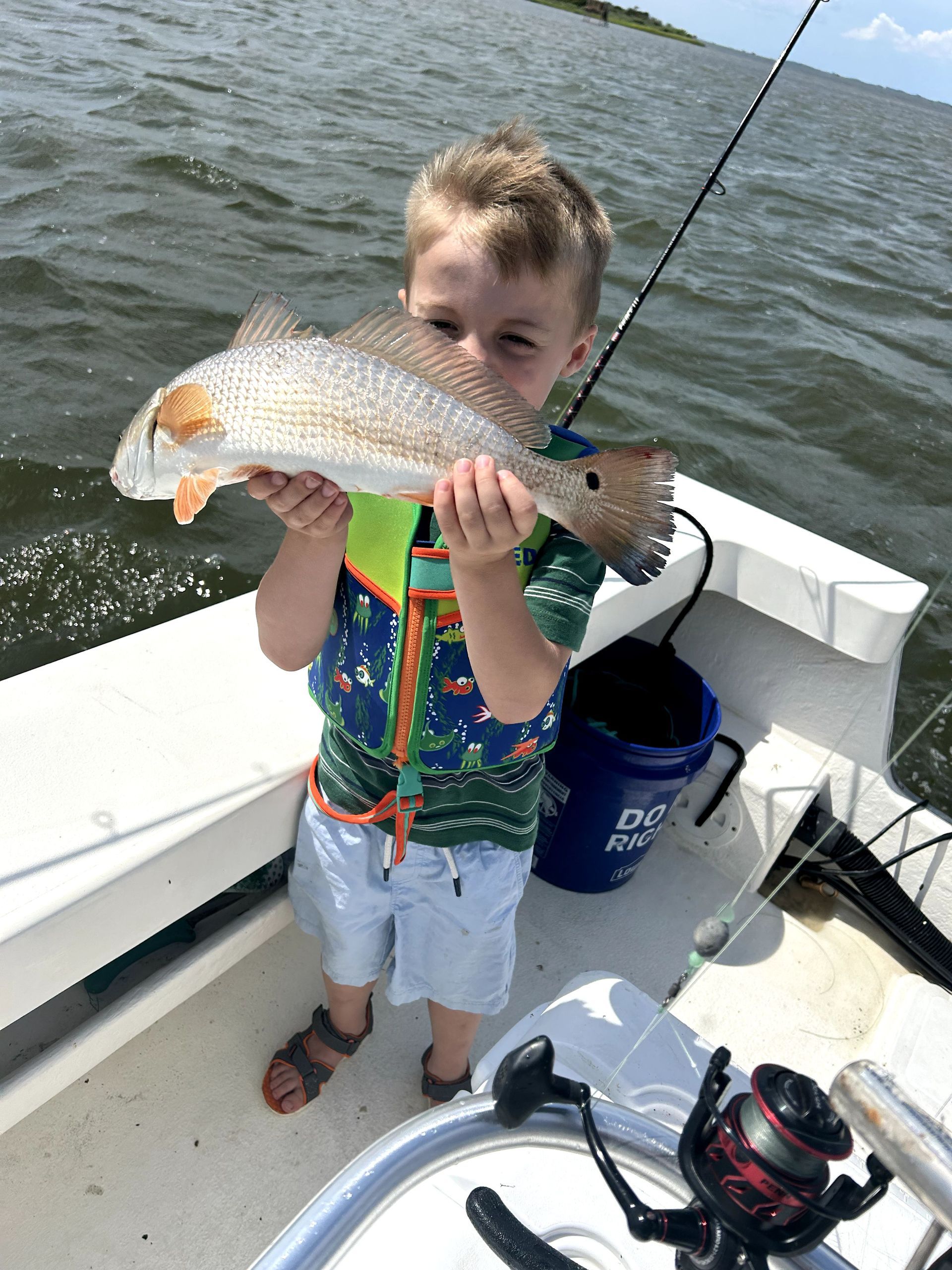 Child holding a large fish on a boat, with fishing gear and water in the background.