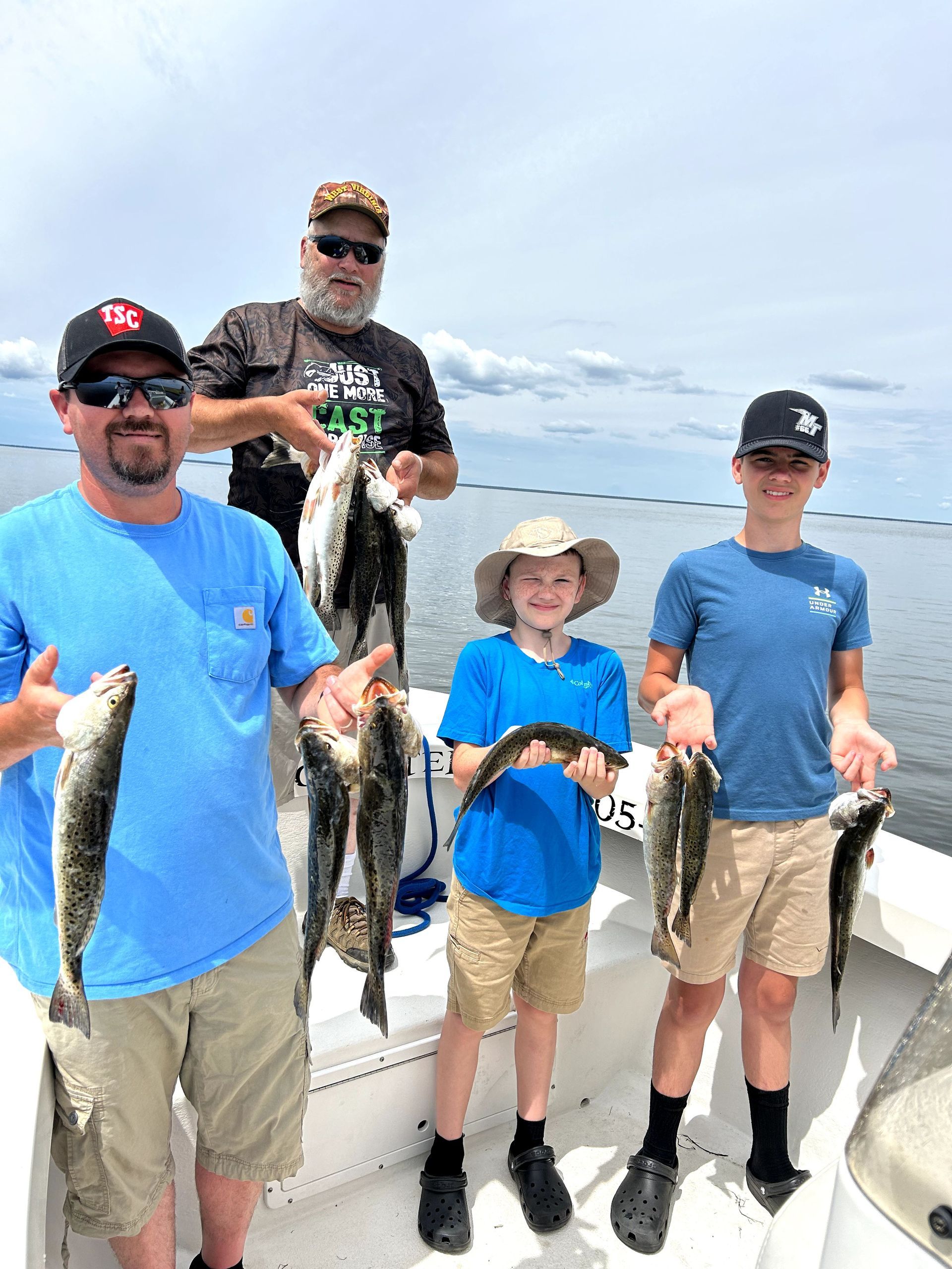 Four people on a boat proudly holding several fish against a bright waterfront sky.