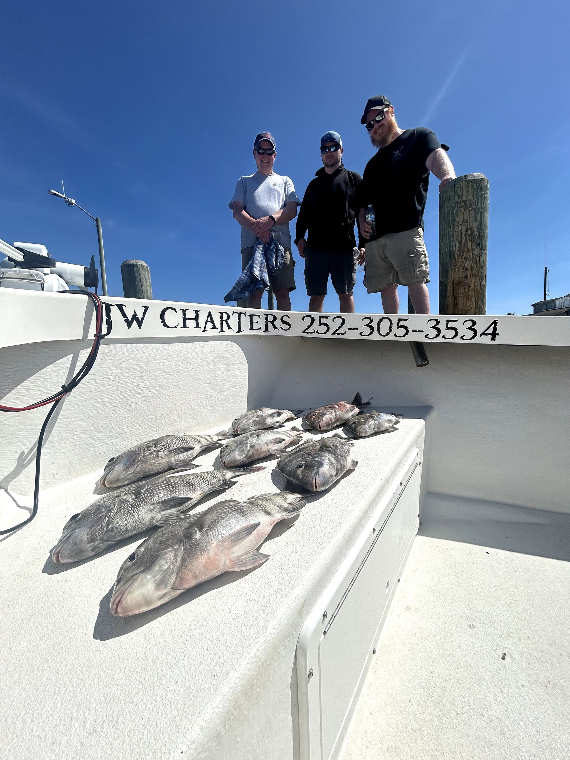 Three people on a boat beside a pile of fish, with a charter boat name visible on the rail.