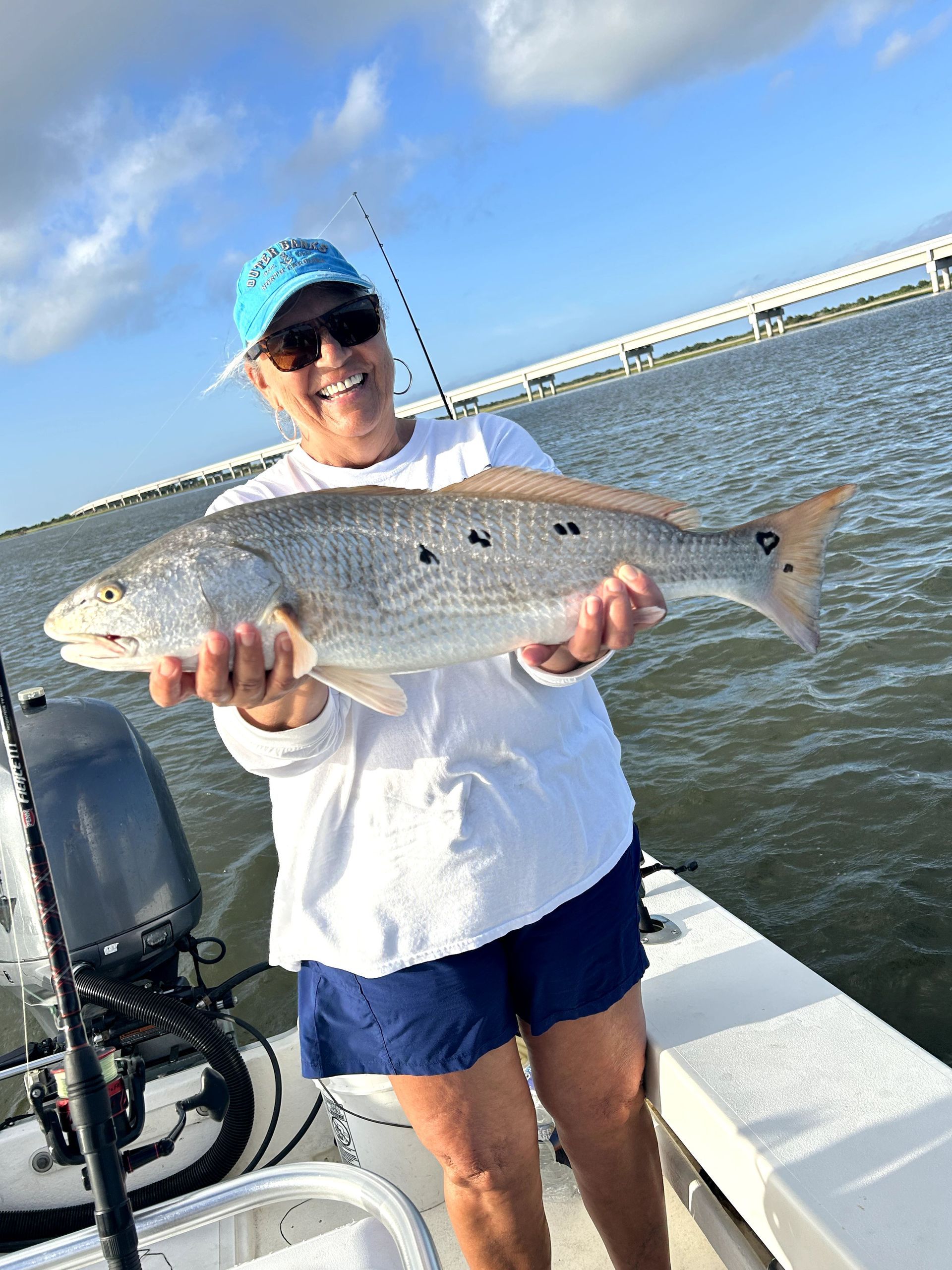 Person holding a large fish on a boat by the water, wearing a blue cap and sunglasses