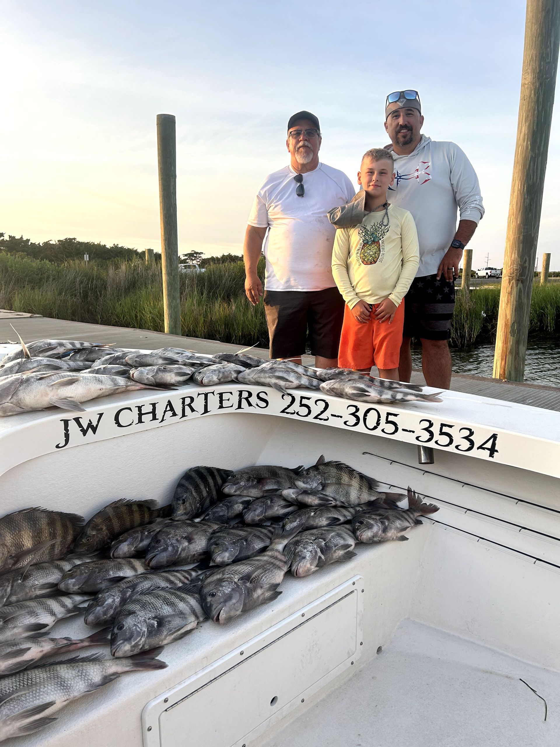 Three people posing on a fishing boat with a large catch of fish in the foreground at sunset.