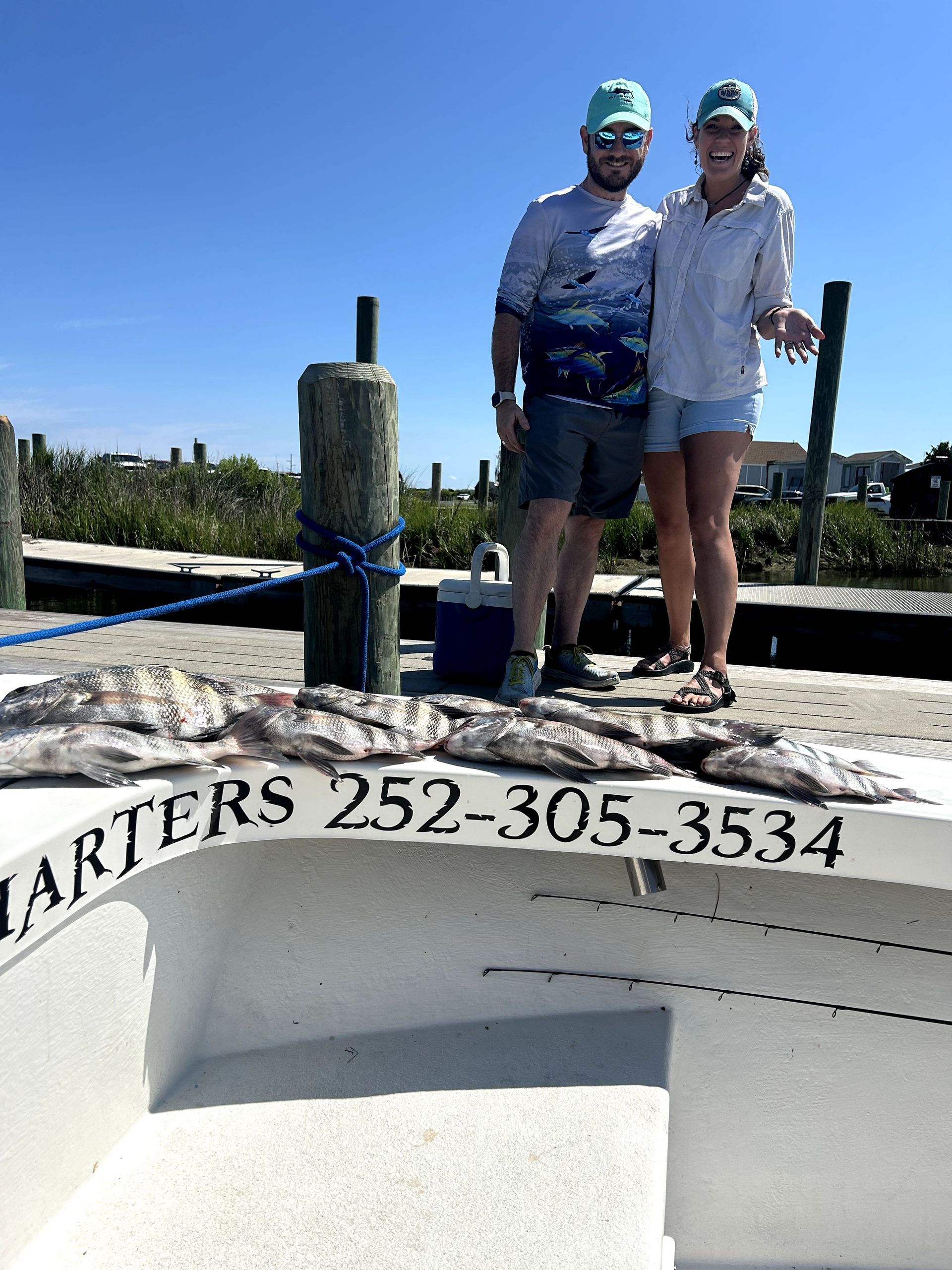 Two people standing on a boat with “QUARTERS 252-305-3534” painted on the bow under a blue sky