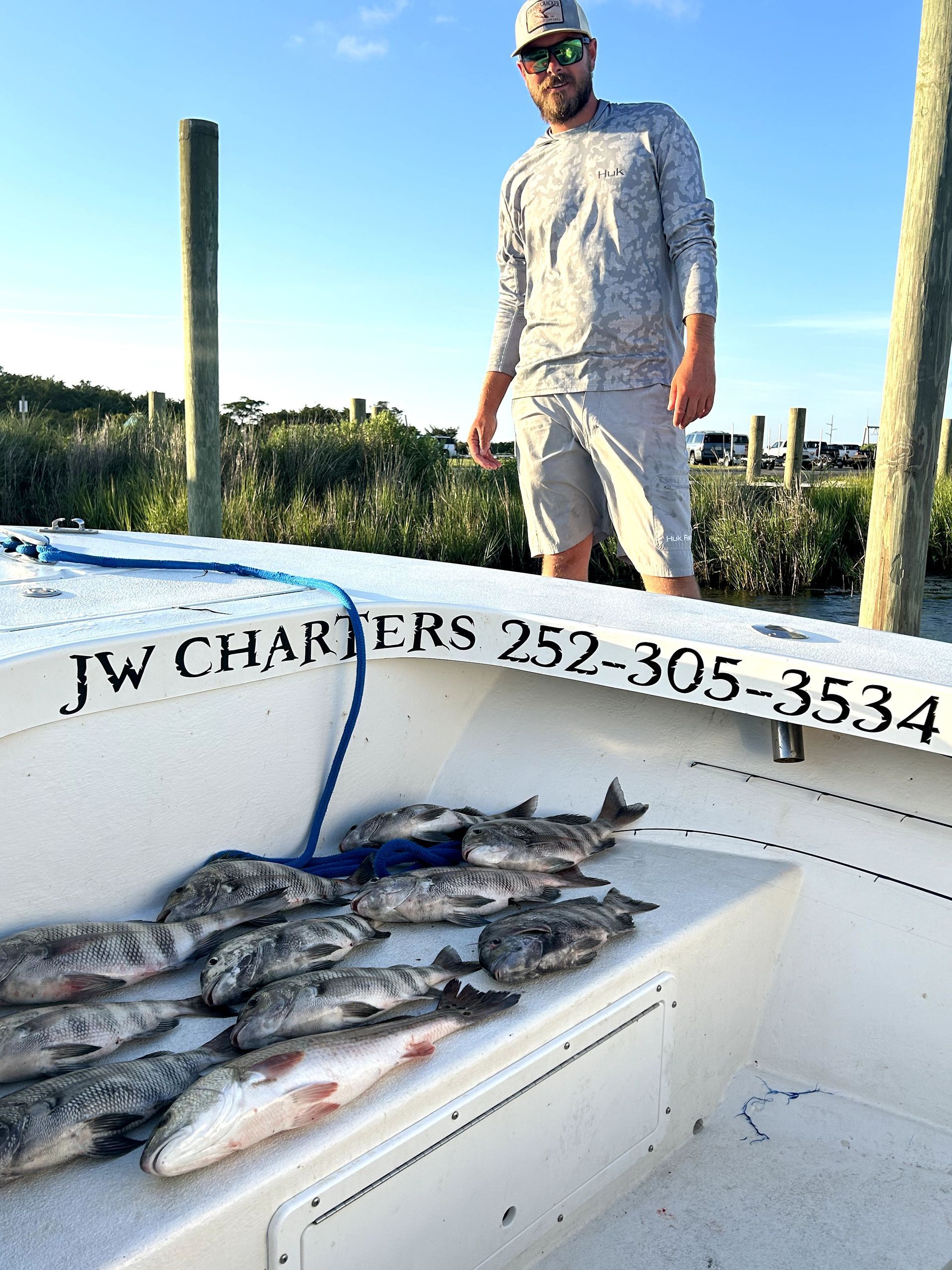 Man standing on a fishing boat with several caught fish on deck, dock posts and water behind him.