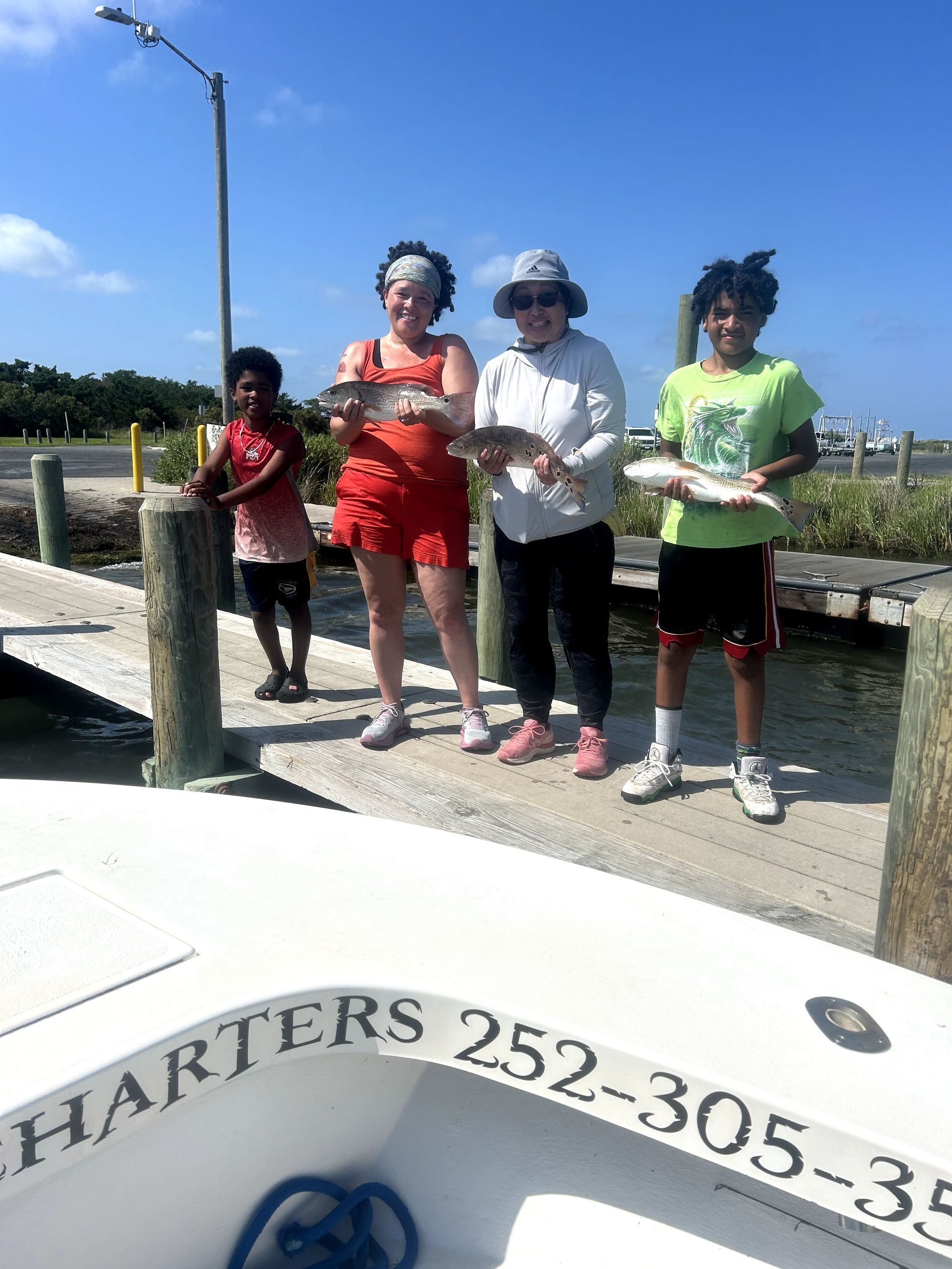 Four people stand on a dock holding fish, with water and a boat in the background.