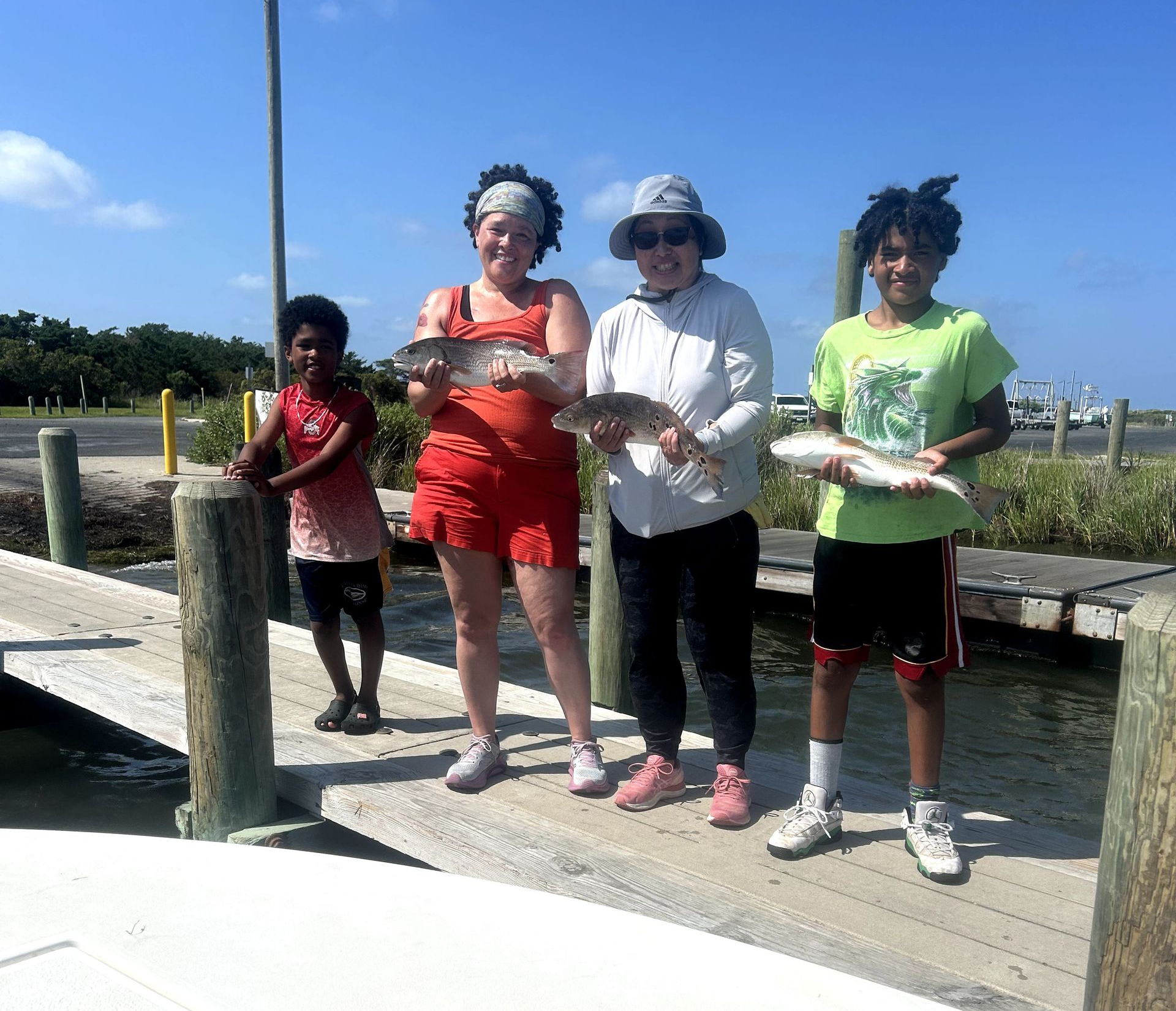 Four people standing on a dock holding fish, with water and a blue sky behind them.