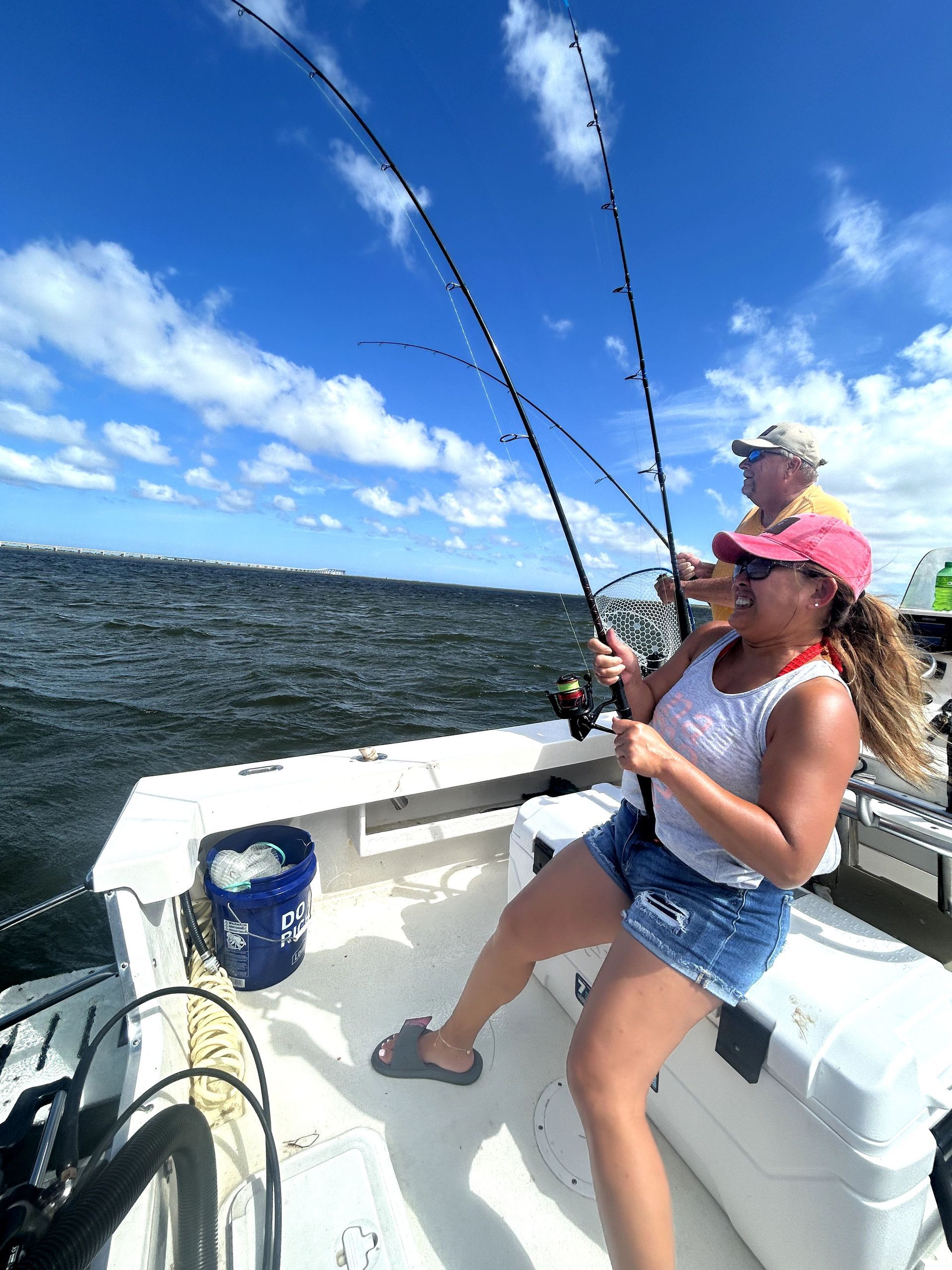 Two people fishing on a boat under a bright blue sky with white clouds.